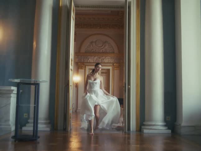 Slow-motion pull-back shot of a graceful bride in a flowing white gown walking barefoot through a grand palace ballroom