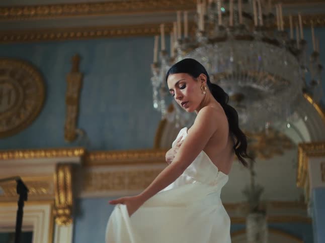 A graceful woman in a flowing white gown dances in slow motion inside an opulent ballroom with a crystal chandelier