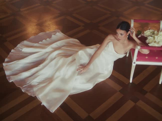 An elegant bride in a beautiful white gown reclines on a polished parquet floor next to an ornate chair with her wedding bouquet