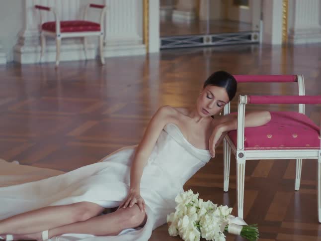 A stunning bride in a white gown reclines gracefully on the floor of a classic ballroom, her bouquet resting beside her