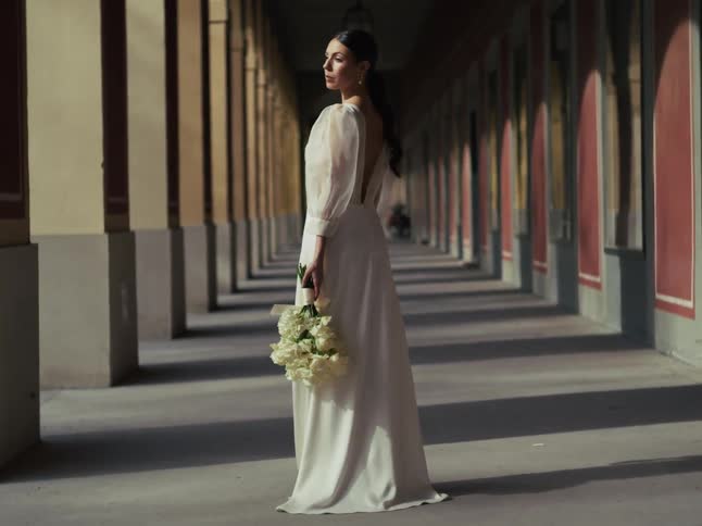 A graceful bride in a white wedding dress holding a bouquet of roses poses elegantly in a long, sun-dappled architectural colonnade