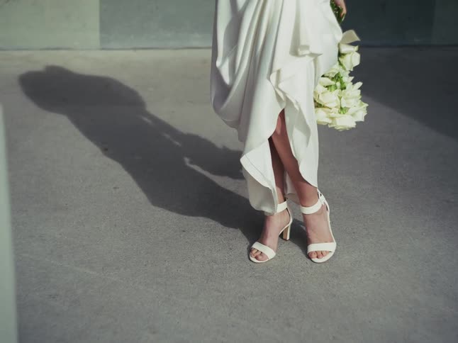 A bride in a stylish white dress and heels holds a bouquet of white roses, casting a strong shadow on the concrete