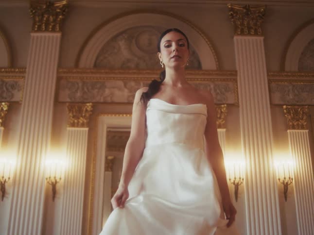 An elegant woman in a beautiful white gown walks gracefully through a grand and opulent ballroom in slow motion