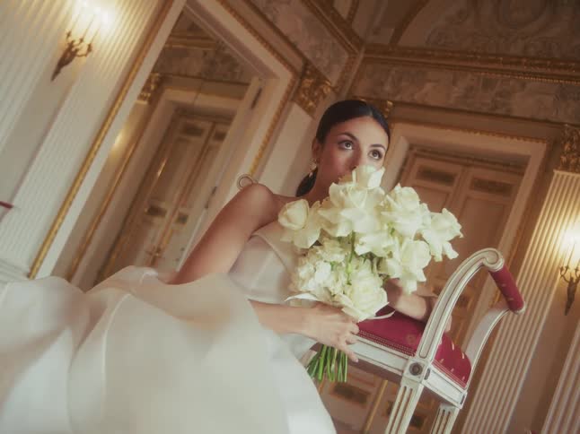 An elegant bride in a beautiful white gown holds a bouquet of white roses as she sits in a luxurious hall, her expression turning from pensive to a joyful smile