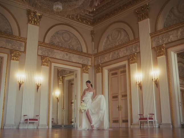 A bride in a white gown poses with a bouquet in a magnificent golden ballroom as her dress flows gracefully in slow motion