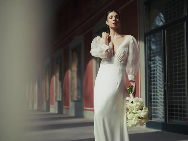A stunning bride poses in her elegant white wedding dress with a bouquet of flowers in a long, sunlit corridor