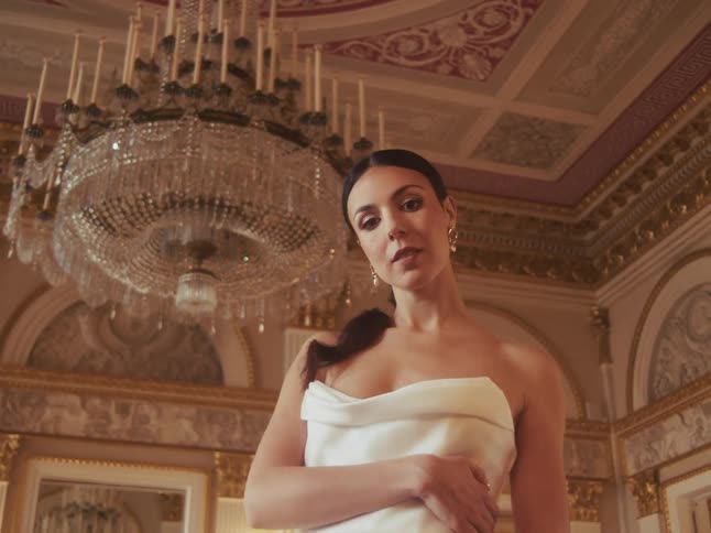 A beautiful woman in an elegant white dress is revealed in a luxurious ballroom, standing beneath a grand crystal chandelier