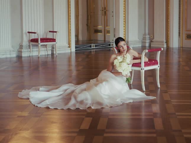 A pensive bride in a flowing white gown sits on the polished floor of an opulent ballroom, leaning against an ornate chair with her wedding bouquet