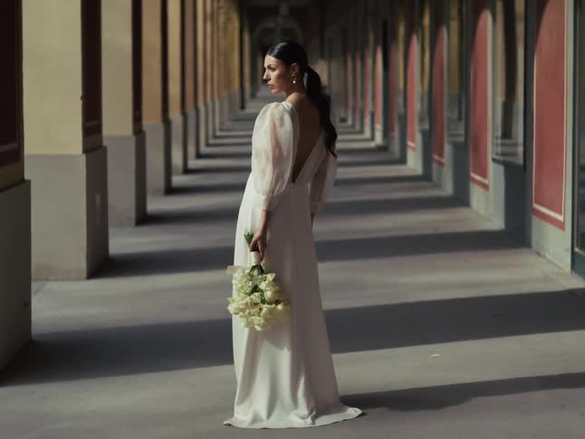 An elegant bride in a beautiful white gown holds a bouquet of roses while standing in a sunlit architectural colonnade