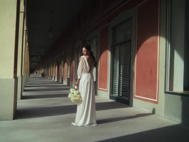 A bride in an elegant white gown holds a bouquet of white roses and poses in a long, classic portico, with dramatic light and shadows creating a beautiful pattern