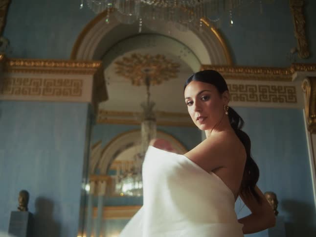 An elegant woman in a lavish room looks over her shoulder as she moves a flowing white sheet to reveal herself to the camera