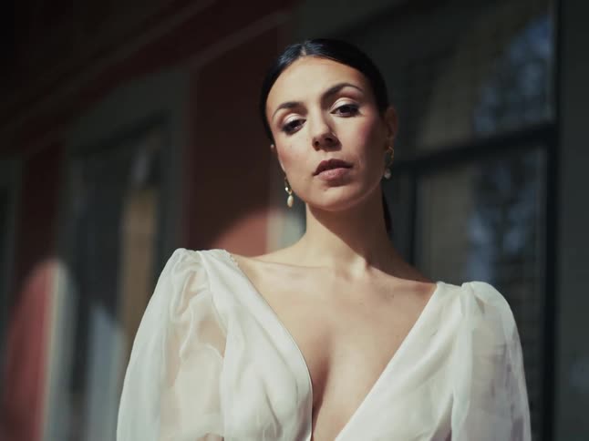 A beautiful dark-haired woman in a sheer white blouse poses elegantly in the sunlight while looking intently at the camera
