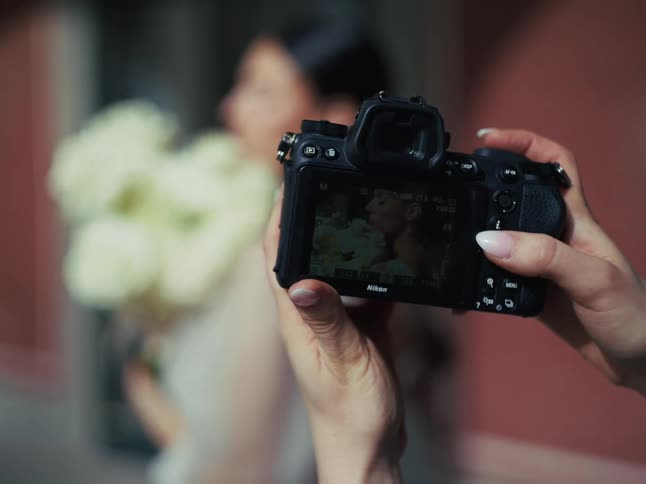 Close-up shot shows a photographer's hands holding a camera and reviewing a portrait of a bride with her bouquet