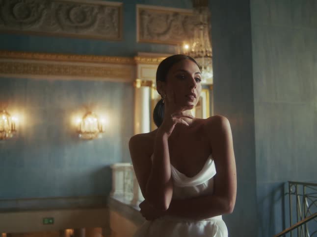 A pensive woman in a beautiful white gown poses elegantly in the dramatic light of a lavish classical building