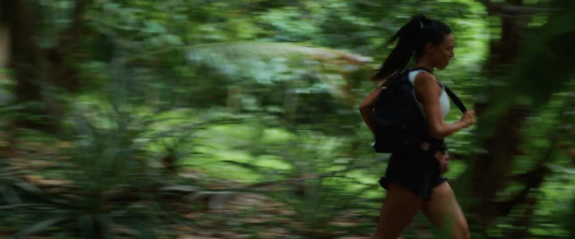 An athletic woman with a backpack urgently runs through a dense tropical jungle