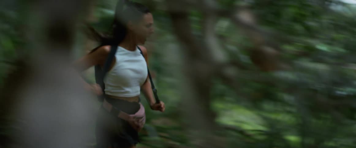 A terrified woman frantically runs through a dense jungle, looking back as if she is being chased