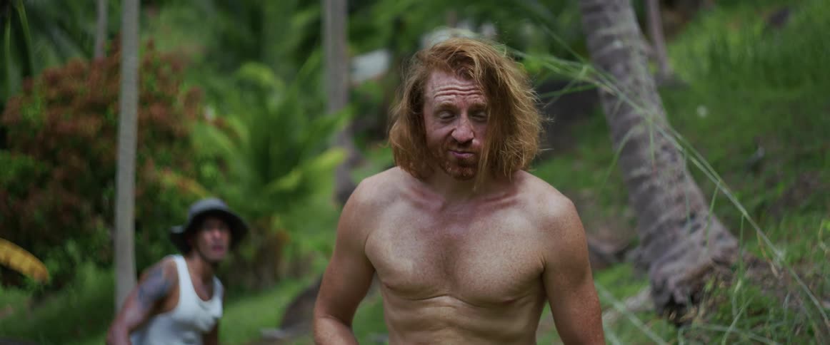 A shirtless man with long red hair stands in the jungle, comically eating a snack and making funny faces