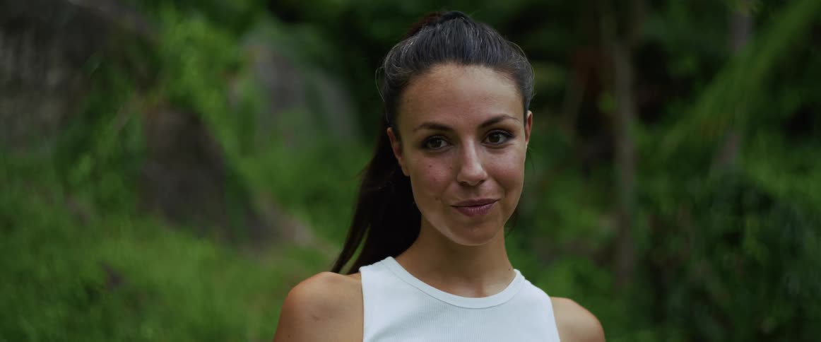 A close-up portrait of a beautiful young woman with her hair in a ponytail smiling warmly in a lush green forest