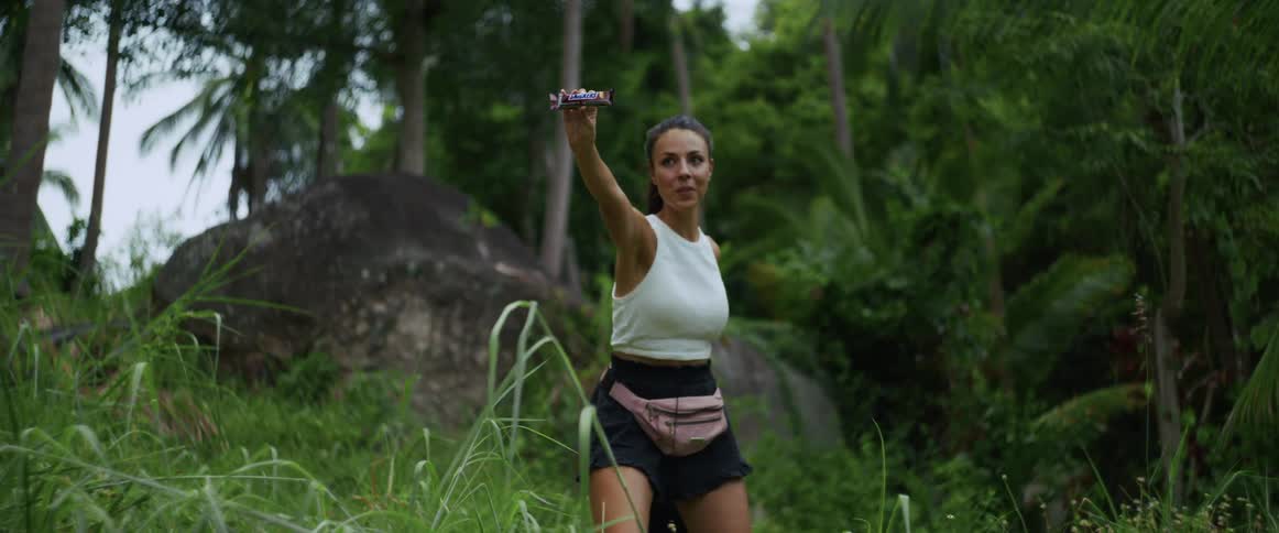 A woman hiking in a lush green jungle pulls out a candy bar and playfully offers it to the camera