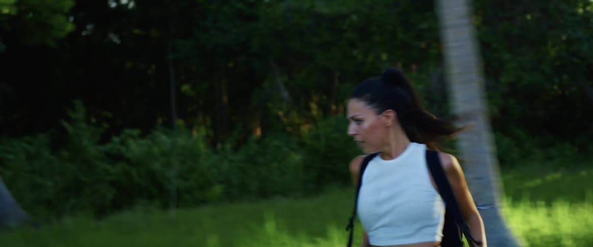 A terrified woman with a backpack runs frantically through a tropical jungle, looking over her shoulder in fear