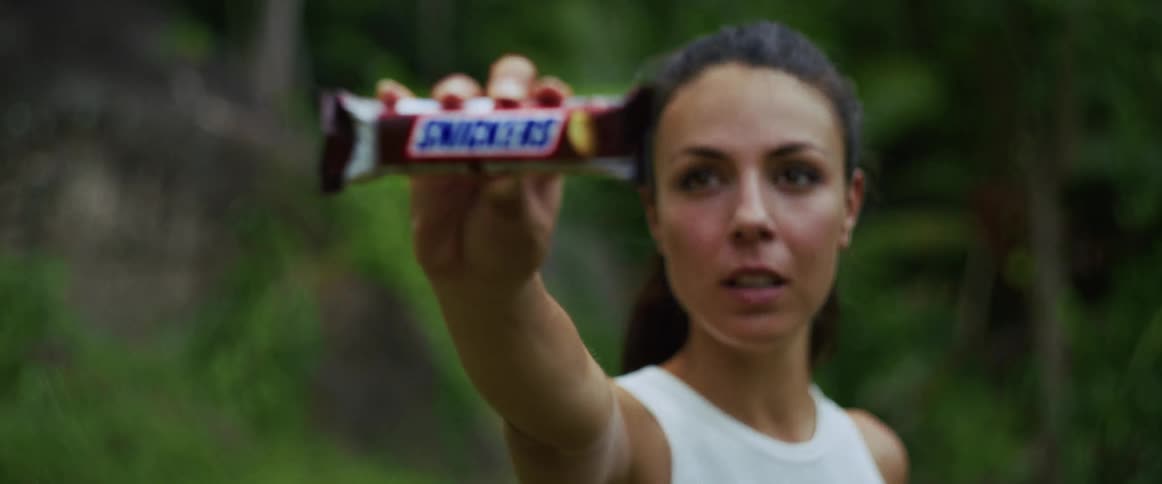 Close-up of an athletic woman holding out a Snickers candy bar in a lush green forest