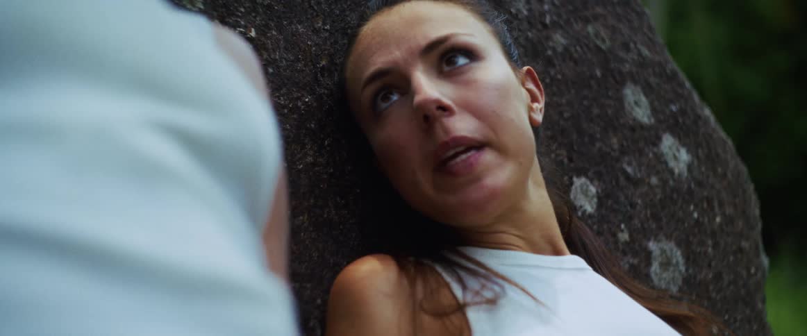 A woman in a white top leans against a giant rock in the forest, breathing heavily and speaking incoherently in a trance-like state