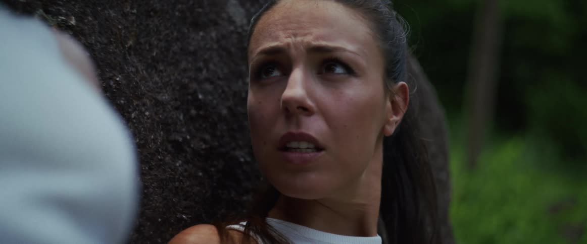 A distressed young woman with her hair in a ponytail gasps for air while leaning against a large rock outdoors