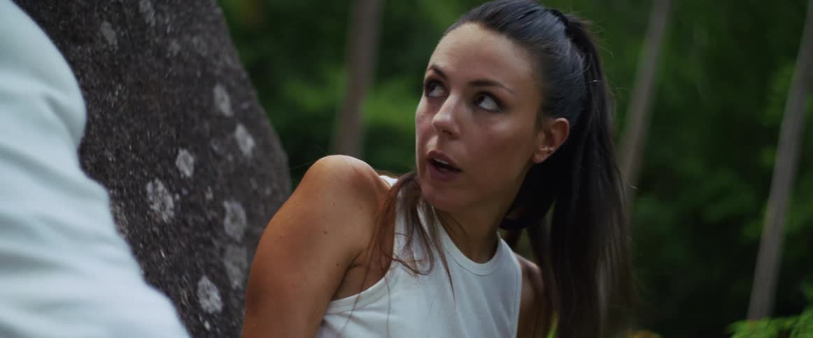 A terrified woman with blood on her face hides in the jungle, breathing heavily and looking around fearfully