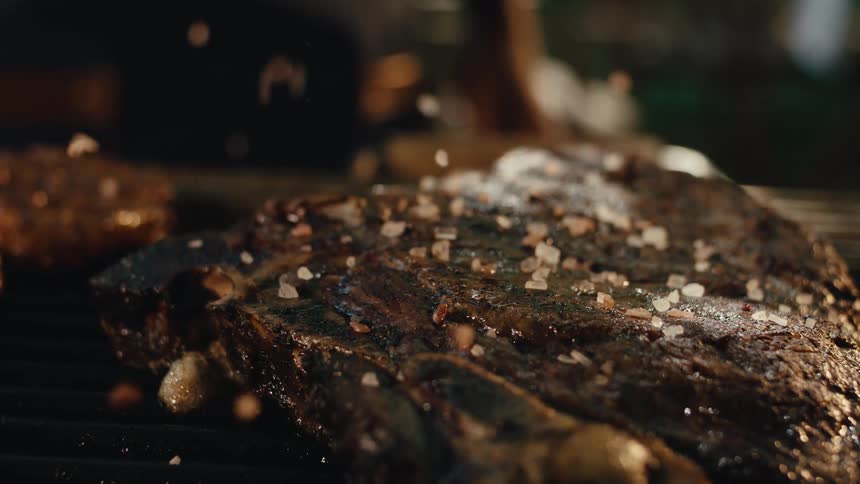 Coarse salt is sprinkled over a juicy bone-in steak on a hot grill in dramatic slow motion