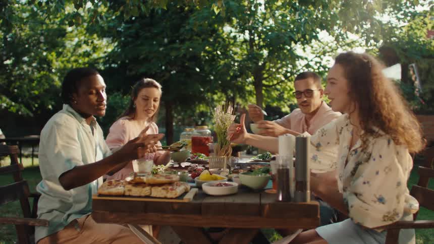 A diverse group of young friends shares a joyful meal together at a wooden table in a sun-drenched backyard garden