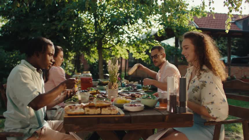A group of diverse young friends enjoys a festive outdoor lunch together at a rustic table in a sunlit garden