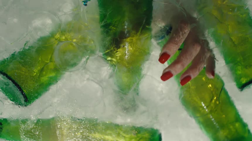 A woman's hand with red painted nails pulls a green glass beer bottle out from a block of melting ice
