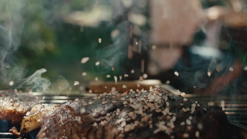 Close-up slow motion shot of coarse salt falling onto a sizzling steak on an outdoor barbecue