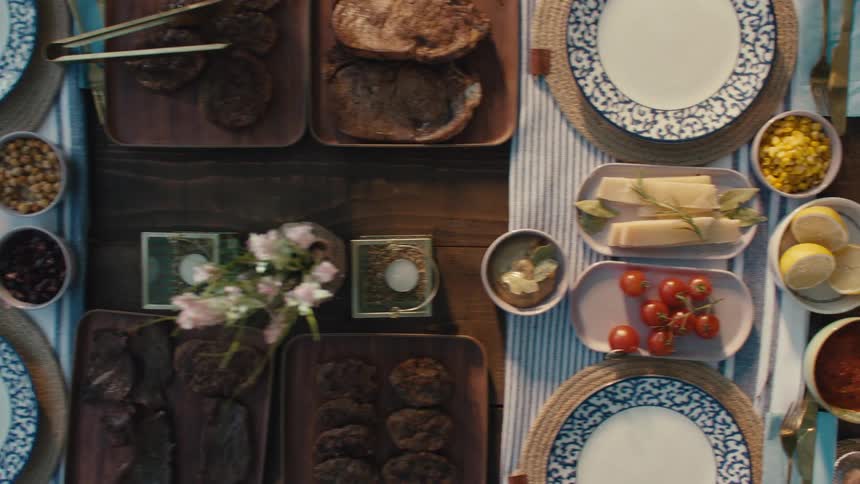 An overhead slow pan reveals a beautifully arranged wooden table being set with a delicious spread of food for an alfresco meal