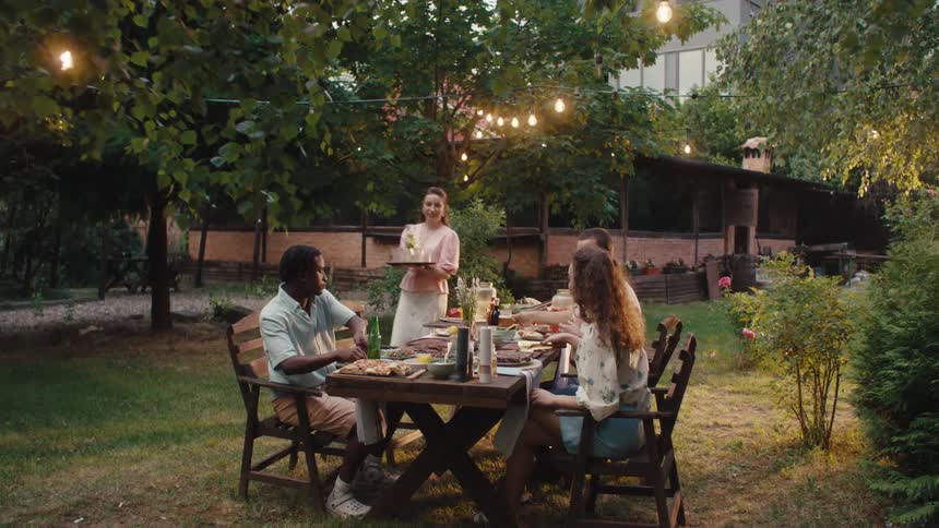 A young woman brings a tray of drinks to her friends who are enjoying an evening dinner party in a beautiful backyard