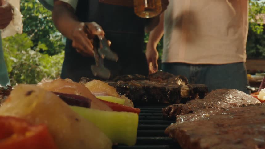 A chef flips a sizzling steak on a crowded barbecue grill as friends watch during a sunny backyard party