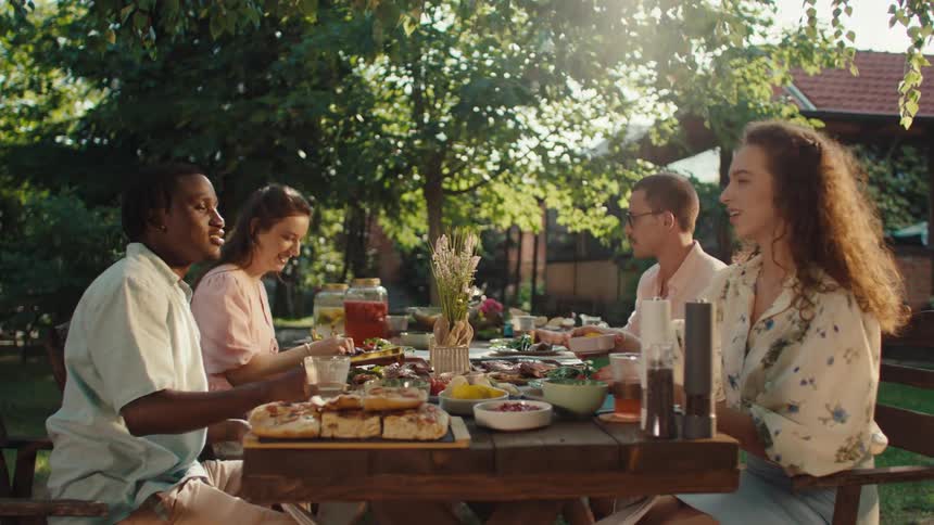 A multicultural group of friends enjoy a leisurely summer lunch together at a rustic table in a sunlit garden