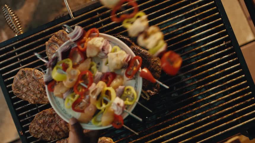 A top-down slow-motion view of a chef grilling a juicy T-bone steak, burgers, and vegetable skewers on a charcoal barbecue