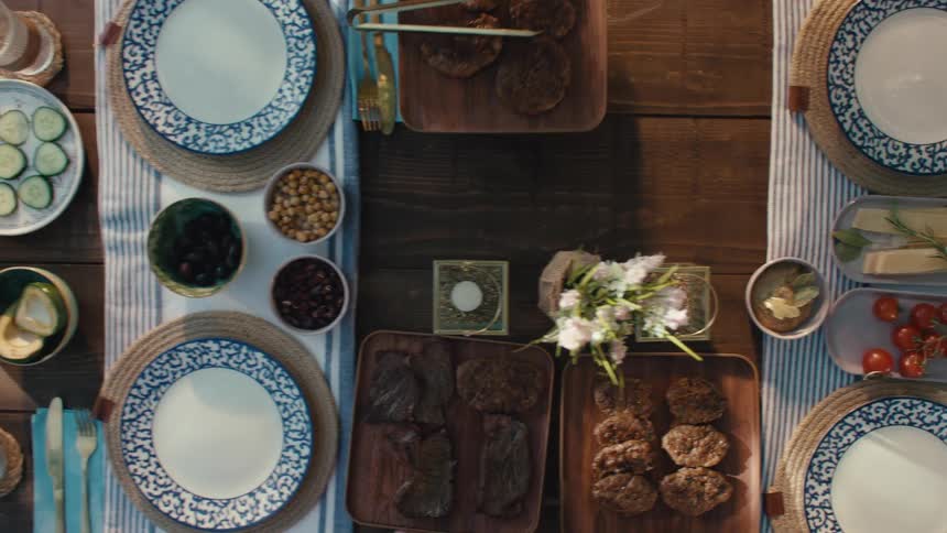 Top down shot reveals a rustic wooden table being set with a delicious and colorful feast for an outdoor dinner party