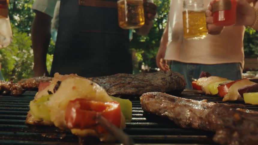 A man flips a large piece of meat on a barbecue grill while friends toast with drinks in the background during a sunny backyard party