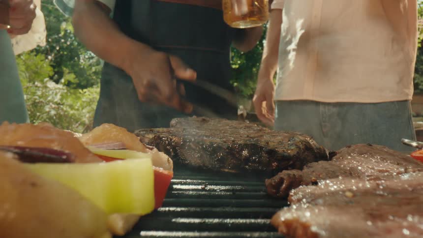 An extreme close-up shot pulls back to reveal a person flipping steaks and veggie skewers on a smoking barbecue grill during a sunny backyard party
