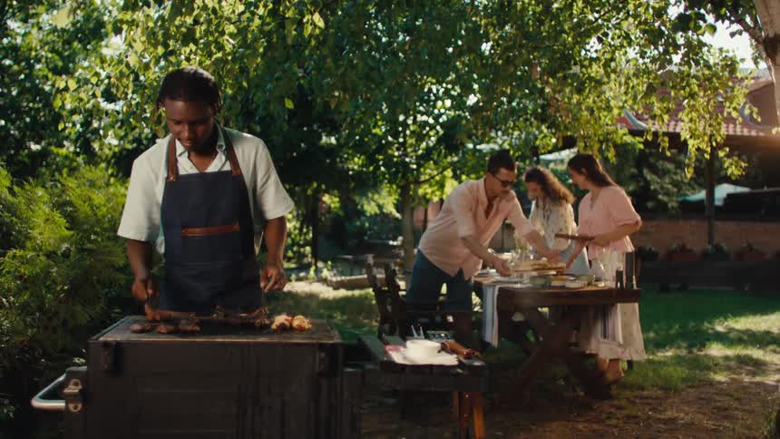 A young man grills meat on a barbecue for his friends as they set the table for a party in a sunny backyard