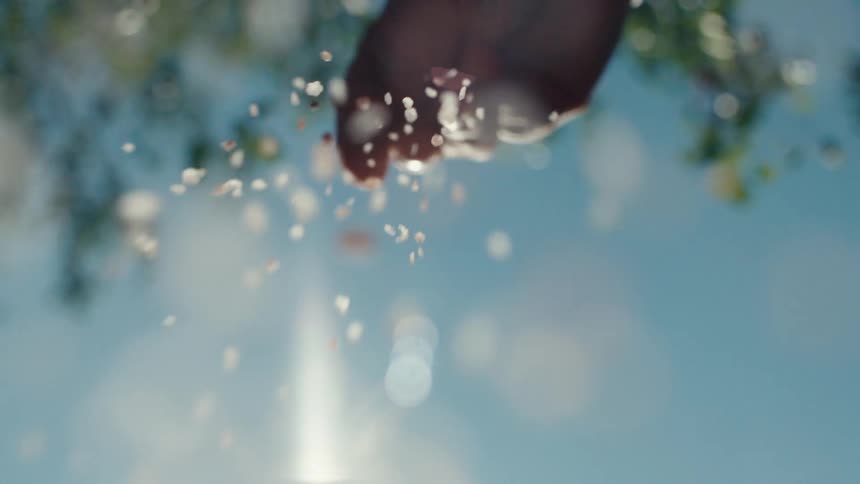 A low-angle, slow-motion shot of a silhouetted hand sprinkling grains of salt against a bright blue sky with a brilliant sun flare