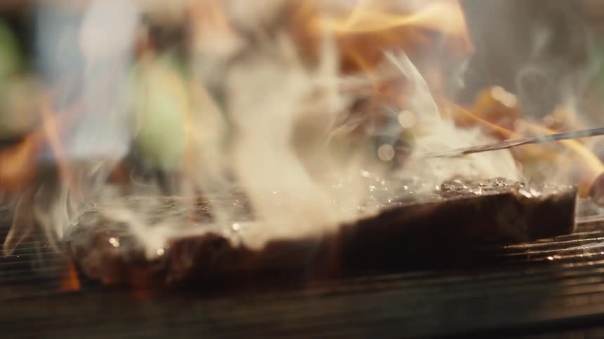In a dramatic slow-motion close-up, a seasoned steak is placed on a hot barbecue grill, sizzling and becoming engulfed in flames before being flipped
