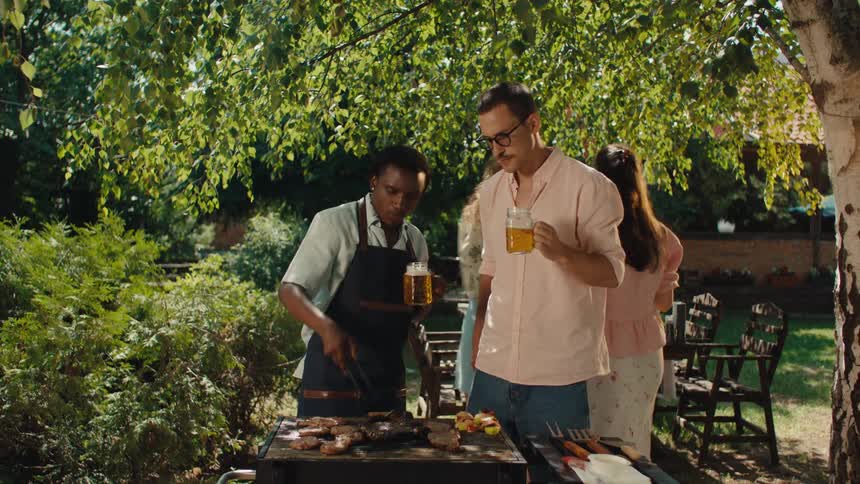 Diverse friends toast with beers and enjoy grilling meat at a sunny backyard barbecue party