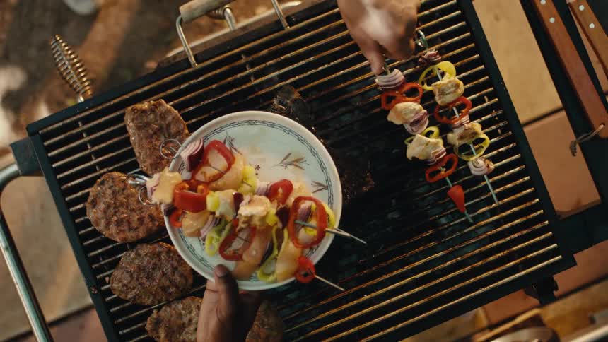 Top-down slow-motion view of a juicy T-bone steak, burgers, and colorful vegetable skewers being grilled over hot charcoal