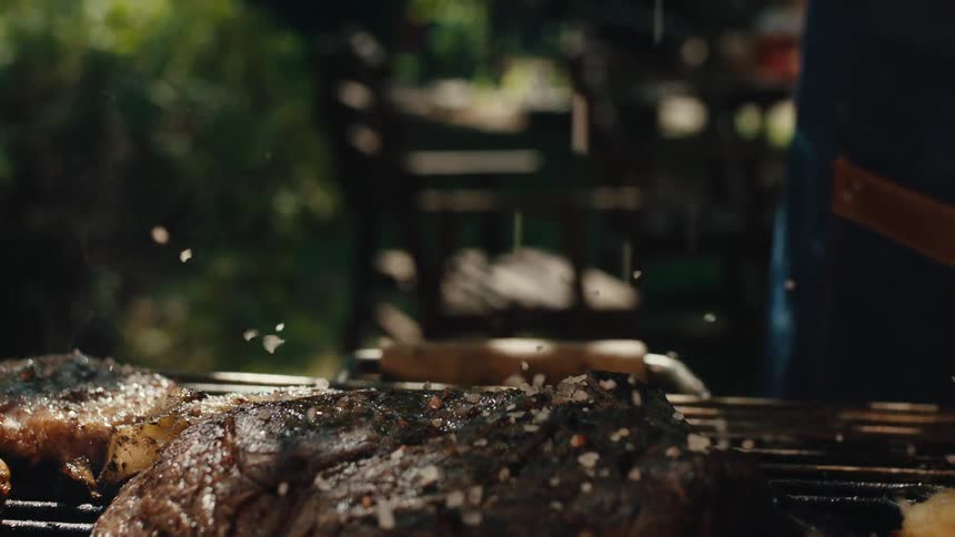 A slow motion close-up of coarse sea salt being sprinkled over a steak, burgers, and vegetable skewers sizzling on a hot barbecue grill