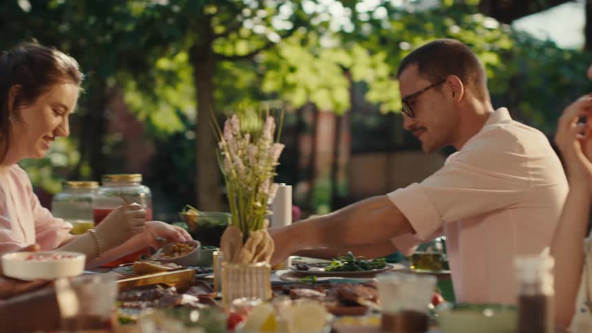 A diverse group of young friends share a meal and conversation at an outdoor garden party on a sunny day