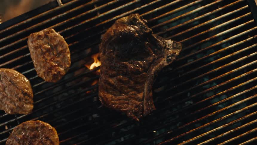 Top-down slow-motion view of a juicy T-bone steak, burgers, and vegetable skewers cooking on a flaming barbecue grill