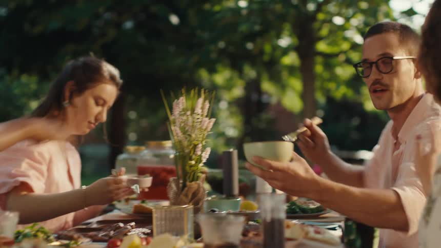 Diverse friends share a joyful meal together at a beautifully set table in a sun-drenched garden, passing food and engaging in lively conversation